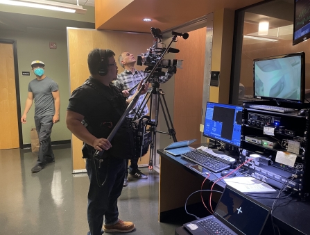 A small film crew points a camera off screen. They are standing in a room with lots of computer screens.