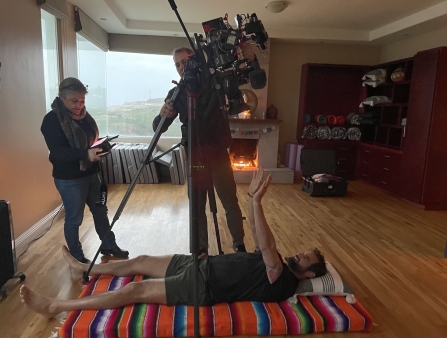 In a beige empty living room, two filmmakers are recording a man laying on his back on a colorful blanket.