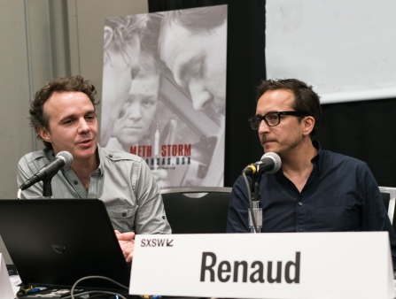 Two men with short brown hair sit at a table with the name tag "Renaud."