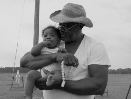 A Black man wearing a straw hat holds a baby.