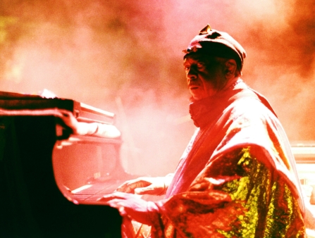 A Black amn wearing an elaborate shiny cape sits at a piano during a concert.