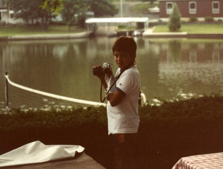 A young boy holds a camera while standing in front of a lake.
