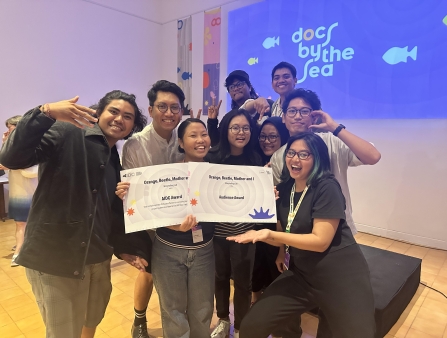 A group of smiling young Asian festival attendees pose with two award certificates against a Docs by the Sea display