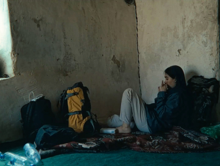 In the corner of a room, a young woman sits next a stuffed backpacked.