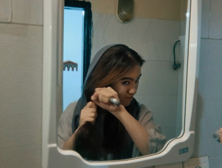 In the mirror's reflection, a young woman uses a knife to cut off her hair.