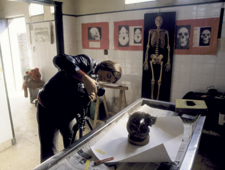 A woman adjusts a tripod camera in front of a skull, in a white-tiled room with various skeleton images