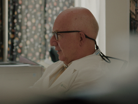 A bald man wearing glasses and a doctor's coat sits in his office