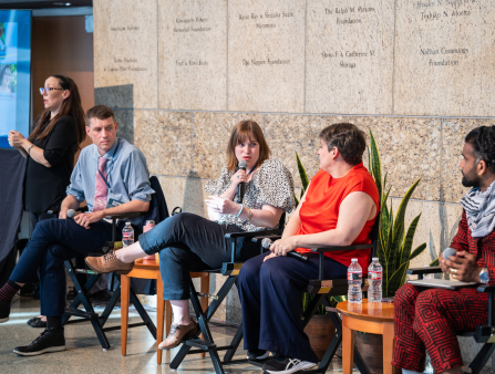 Four people sit in chairs in a bright large hall. There is an ASL interpreter in the far left.