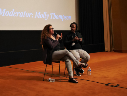 On a carpeted stage, two people in black tops and gray pants sit on chairs.