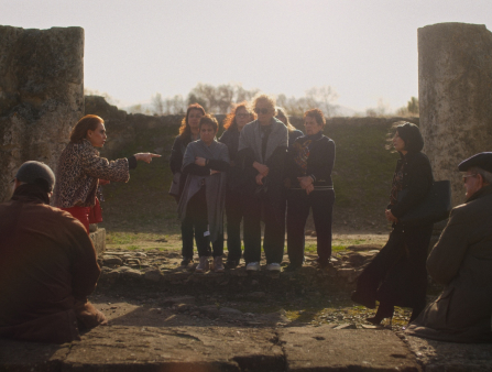 Two women on opposite sides of an outdoor amphiteatre stand aggressively staring at each other, while a group between (and some others seated on either side of them) observe them.