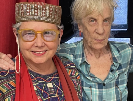 Two older white women, one wearing glasses, the other a checkered shirt, smile and pose together