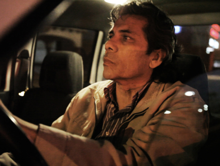 A brown-skinned man with dark medium-length hair, lit by street lights at night, is at the wheel in his car