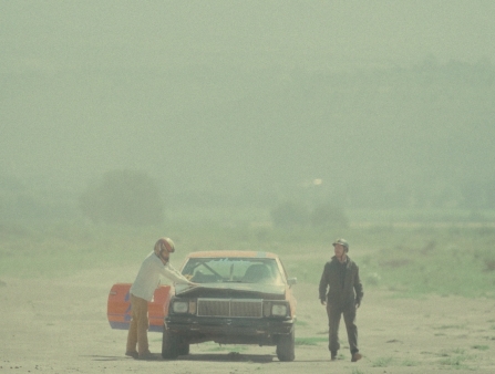 In a dusty road, two men with helmets on stand on either side of a broken down, battered car.