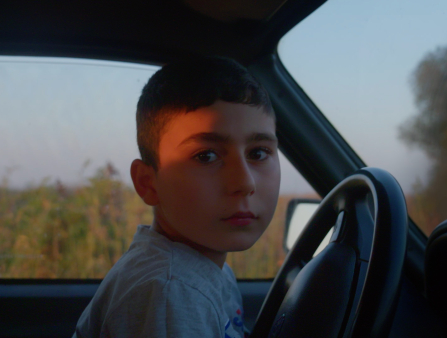 A teenaged boy sits in a car, looking at the camera.