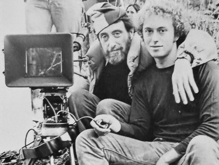 Black and white image of two men, one bearded, one clean-shaven, sitting down outdoors next to a film camera.