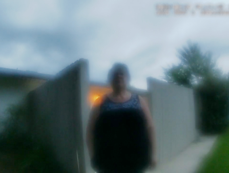 Blurry bodycam shot of a white woman wearing a tank top standing in front of a fenced yard.