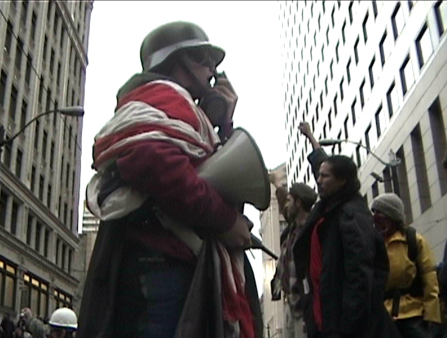 A man draped in the American flag with a protective helmet and glasses, using a megaphone at an outdoor city protest