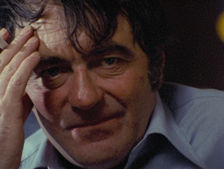 Tight close-up of a white man with dark hair, hand on his forehead, looking straight at the camera