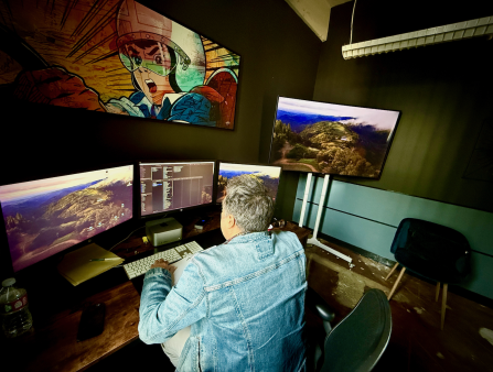 A man with grey hair and a denim jacket is seen from behind as he works at an editing bay, a poster of Speed Racer right above his workstation