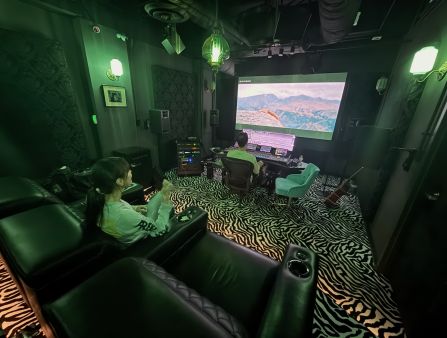 A white woman with long black hair in a ponytail looks at a man working on a big screen in front of him at a green-lit office with zebra-print carpet