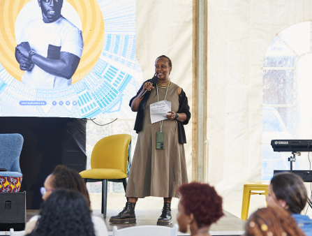 A Black woman in a simple long grey dress with a black sweater, stands on a stage with a mic addressing a gathered crowd