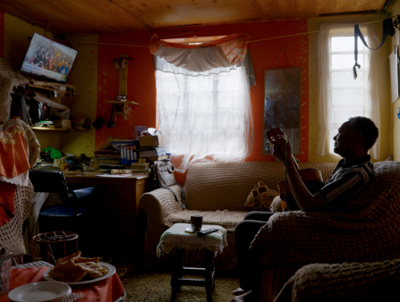 An older Black man plays a guitar while sitting in a crowded living room as he watches a small TV mounted on the wall.