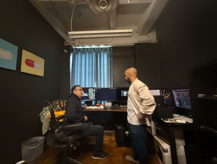 Two men, one sitting, another standing, both wearing casual clothes, talk at a darkened office full of screens