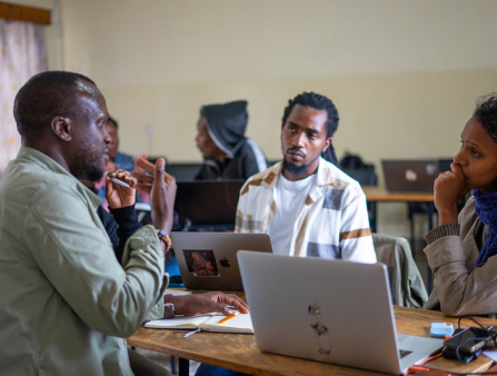 A Black man in a light shirt sits across from a Black man and a Black woman, both in casual clothes, over a desk with a laptop