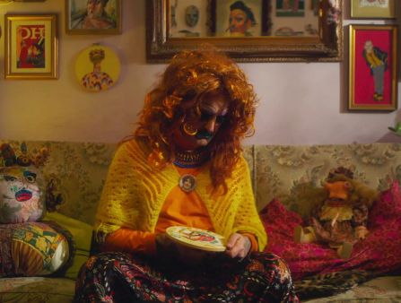 A drag queen wearing a bright orange wig, bold blue lips, and similarly brightly-colored clothes, sits in a cluttered living room sewing