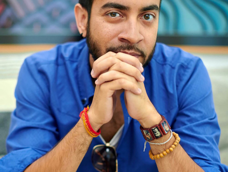 Headshot of Brown man with black hair and a beard, in a bright blue shirt, holding his crossed hands over his face.
