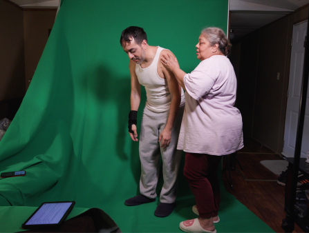 Against a screen screen, an older heavyset Brown woman with white hair in a bun helps a younger brown man with dark hair and a beard in sweats and a tank top keep his posture