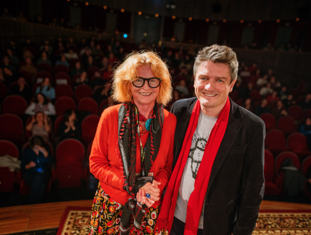 An older white woman with bright red hair, wearing dark-rimmed glasses and scarves with a red cardigan smiles next to a white man in a t-shirt, red scarf and a jacket, in front of a theater audience behind them