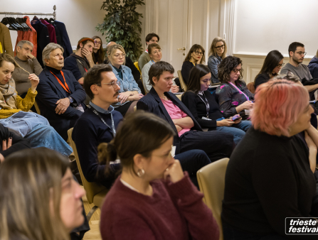 A crowd wearing casual clothing and lanyards sit around in a conference room facing the front of the room