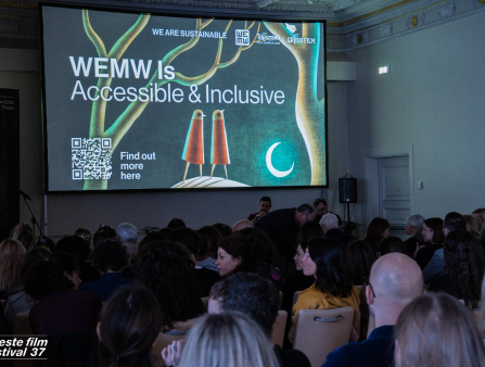 A screen reads "WEMW is accesible and inclusive" in front of a crowd seated in conference chairs in a small enclosed room