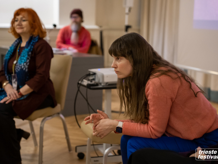 A white woman with long brown hair wears a light orange long-sleeved shirt and a blue skirt; she's seated with a projector behind her; an older white woman with shoulder-length orange hair sits in the background
