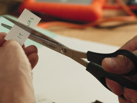 Two light-skinned hands hold a pair of scissors cutting a small piece of paper that reads "It was"