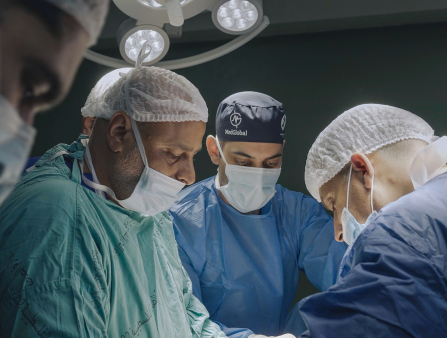 Four brown-skinned doctors wearing surgery scrubs and masks hover over presumably an operating table
