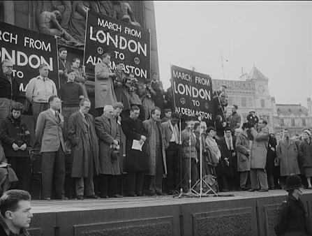 Black and white image of men and women in coats standing in front of microphones with protest signs that read "March from London to Aldermaston"