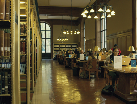 A pristine-looking library is lit by a big window to the right, with stacks of books seen on the left as a handful of readers and scholars crowd the tables by the window