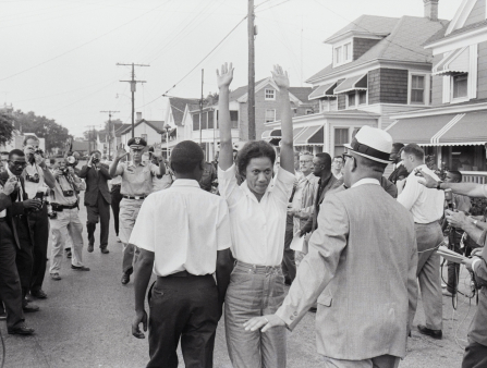 Black and white shot of a Black woman in a casual pants and white shirt holding her hands up while surrounded by protestors out in the streets