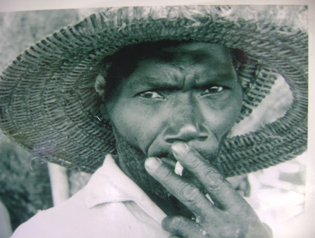 A close-up, black and white photo of a dark-skinned Black man, in a white shirt and wide-brimmed straw hat staring at the camera as he takes a drag from his cigarette