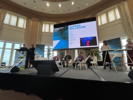 On a large raised stage, two women stand at a podium. Behind them are a row of seated people and a large screen with the information for the "Queens Ballroom" project.
