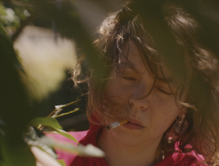 A close up of a white woman with shoulder-length dark hair in a red shirt, eyes closed, holding a cigarette in her mouth outside