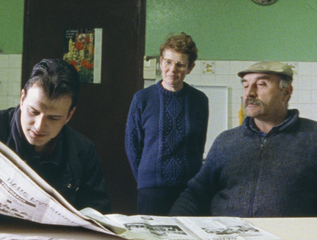 A young white man with dark hair sits at the kitchen table reading the newspaper as an older man with a mustache and hat, and a woman with short curly hair and glasses look on