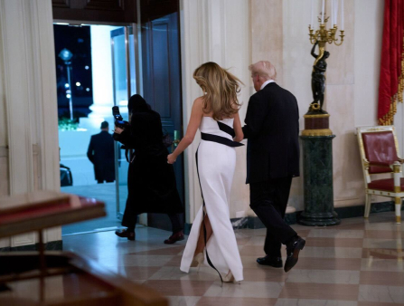 A white slender woman in a black and white dress walks away holding hands with an aging white-haired white man in a dark suit