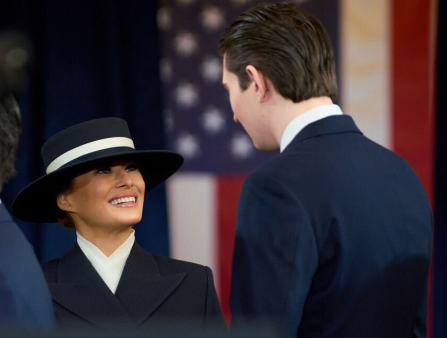 A smiling white woman with a large black and white hat and black and white dress greets a tall white man in a suit with a US flag behind them