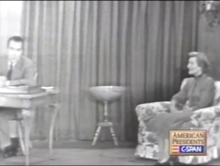 Black and white blurry C-SPAN screenshot of a white man in a suit seated at a desk across from a white woman in a dress sitting at a reading chair