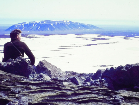 A white man sits on a rock formation on a glacier, a snow-capped mountain is seen in the background