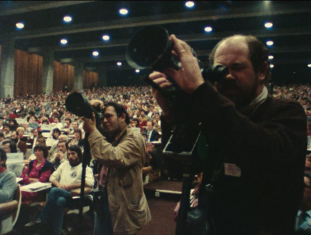 Two male photographers are seen in the foreground against a packed theater behind them
