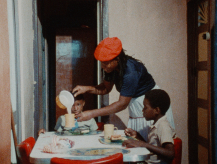 A Black young woman wearing a blue short-sleeved shirt, a white skirt and a bright red orange beret is serving milk at a table to her two young seated boys